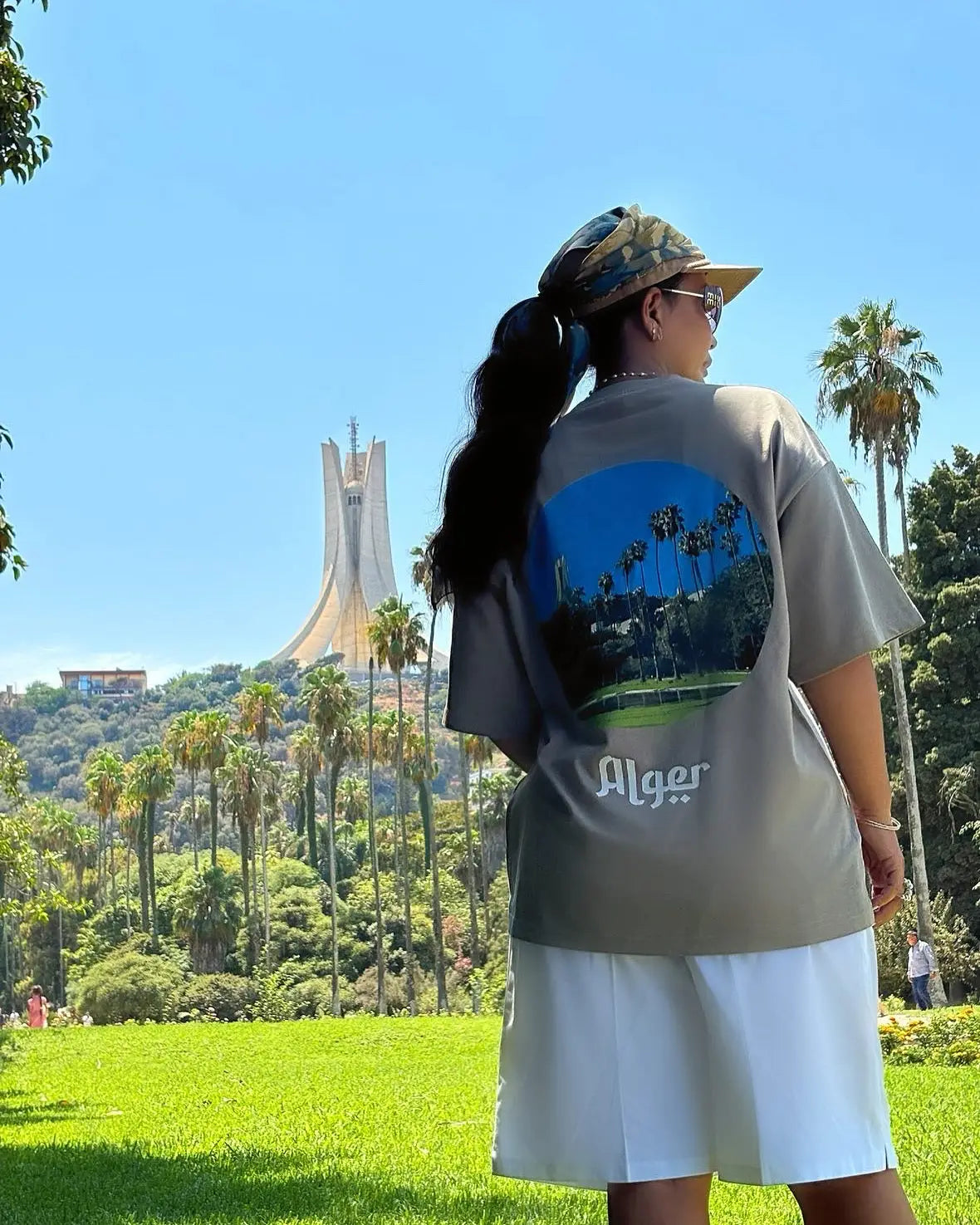 Une personne portant un tee-shirt gris avec un motif circulaire bleu et vert et l’inscription ’Alger’ en dessous, associé à un short blanc.